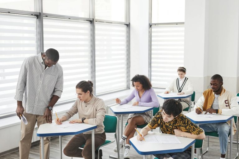A diverse group of students taking a test in a classroom with a teacher assisting.