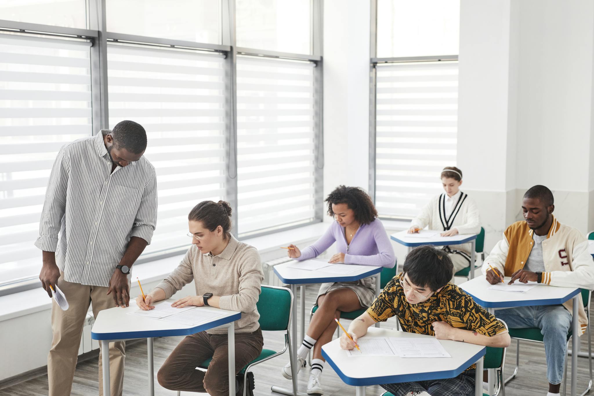 A diverse group of students taking a test in a classroom with a teacher assisting.