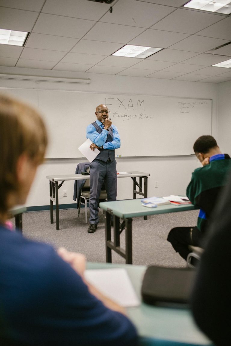 A professor stands attentively in a classroom observing students taking an exam.